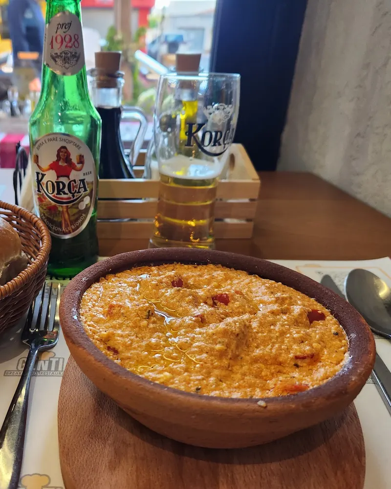 A baked dish in a clay pot, with a basket of bread and a Korça beer